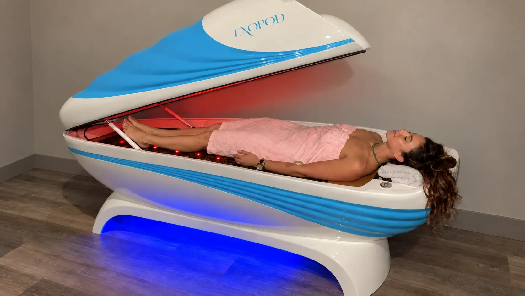 Woman wrapped in a pink towel lying in the white and blue ExoPod with the lid raised and red lights on