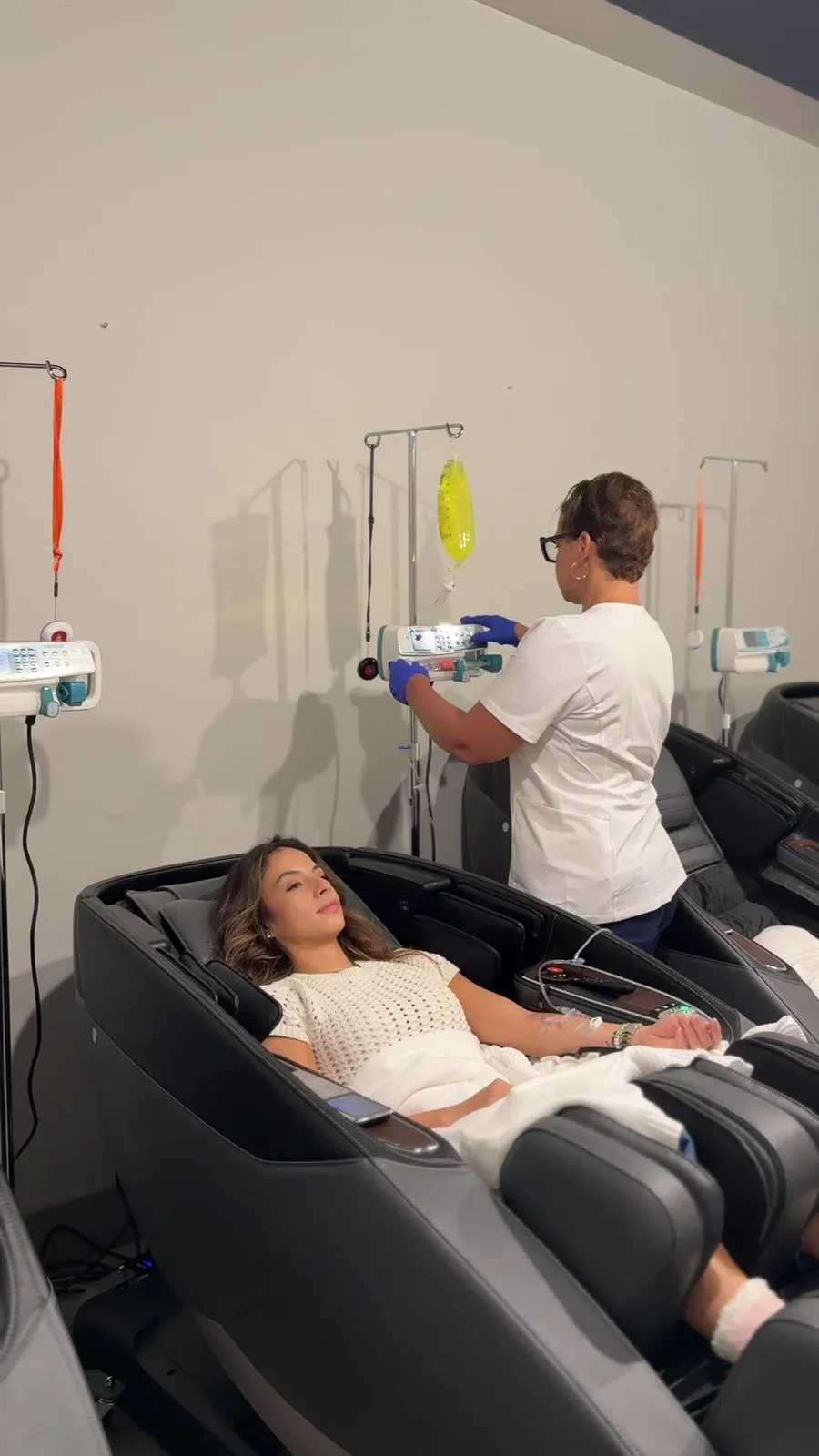 Client reclining in a black treatment chair with an IV line in her arm while a clinician adjusts the infusion pump
